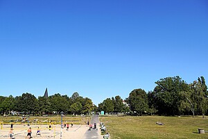 Volleyballfeld auf Sand mit Spielern im Vordergrund, umgeben von Rasenflächen, auf denen Leute liegen oder spazieren. Ein Weg führt durch den Park, Bäume und ein Kirchturm ragen im Hintergrund in den blauen Himmel.