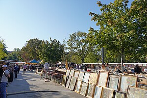 Zahlreiche Gemälde in Bilderrahmen sind an einem Stand ausgestellt, während Besucher durch den Mauerpark-Flohmarkt schlendern. Die Sonne scheint hell und taucht die gesamte Szenerie in ein sommerliches Licht.