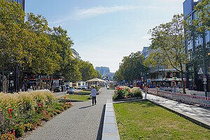 Die Tauentzienstraße in Berlin mit breiten Gehwegen, auf denen Menschen spazieren. Bunte Blumenbeete säumen die Fußwege, während im Hintergrund Geschäfte, Restaurants und dichter Verkehr zu sehen sind. Ein klarer Himmel und grüne Bäume geben der Szene eine lebendige und angenehme Atmosphäre.