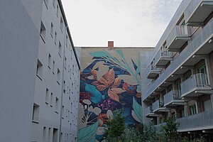 Eine farbenfrohe Wandmalerei mit Pflanzenmotiven in einem Innenhof in Moabit, Berlin. Die Fassade ist von modernen Wohnhausbalkonen eingerahmt.