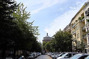 Eine Berliner Straße, gesäumt von geparkten Autos und Altbauten mit Bäumen, die den Himmel teilweise verdecken. Im Hintergrund steht der markante Wasserturm, ein Wahrzeichen des Kollwitzkiezes.