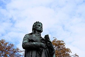 Eine weite Grünfläche im Schillerpark, im Hintergrund die Schiller-Statue und eine Steinmauer mit Bäumen dahinter. Der Himmel ist von großen weißen Wolken bedeckt.