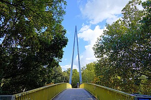 Eine gelbe Fußgängerbrücke mit hohem Bogen im Volkspark Wilmersdorf, umgeben von dichten grünen Bäumen, die den blauen Himmel durchscheinen lassen. Im Hintergrund ist eine Person, die die Brücke überquert.