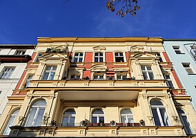 Eine prächtige Altbaufassade mit cremefarbenem Anstrich und reich verzierten Fenstern. Das Gebäude hat schmuckvolle Balkone mit Blumen. Im Vordergrund steht ein Fahrrad angelehnt an eine Hecke, die rote Beeren trägt. Der Himmel ist klar und blau.