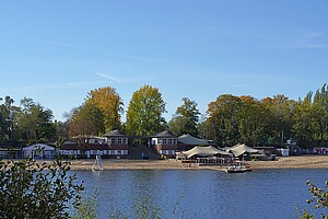 in Blick auf den Plötzensee an einem sonnigen Tag. Der See spiegelt das helle Sonnenlicht wider, während am Ufer grüne Bäume zu sehen sind. Im Hintergrund erkennt man ein paar weiße Gebäude mit Zeltdächern und einer Sandfläche, die an einen Strand erinnert.