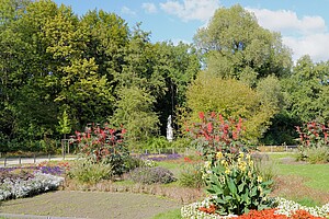 Ein üppig bepflanztes Blumenbeet mit roten, gelben und lilafarbenen Blumen im Vordergrund. Im Hintergrund stehen hohe Bäume, die den Großen Tiergarten in Berlin umrahmen. Eine weiße Statue ist zwischen den Bäumen zu sehen.