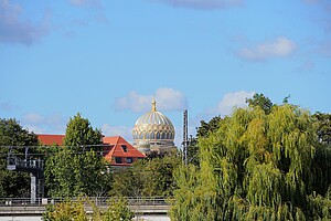 Die goldene Kuppel der Neuen Synagoge ragt über die Dächer Berlins, umgeben von grünen Bäumen und roten Dächern, unter einem strahlend blauen Himmel.