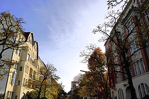 Ein parkähnlicher Platz im Bayerischen Viertel, wo die Statue des Bayrischen Löwen auf einer Säule thront. Der Löwe ist aus grünlich oxidierter Bronze und steht vor kahlen Bäumen in Herbstfarben. Im Hintergrund ragen moderne Wohngebäude mit vielen Balkonen und großen Fenstern auf.