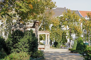 Der Viktoria-Luise-Platz in Berlin mit einer gepflegten Gartenanlage, einem Springbrunnen und umgeben von historischen Wohnhäusern. Im Vordergrund dichter Baumbestand und Hecken, die dem Platz eine idyllische Atmosphäre verleihen.