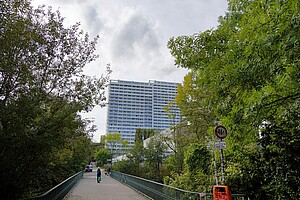 Das Bild zeigt eine Brücke im Fennpfuhl-Park in Berlin, die über einen von Bäumen gesäumten Weg führt. Im Hintergrund erhebt sich ein großer, heller Plattenbau. Eine Person mit einem Einkaufskorb und ein Spaziergänger sind auf der Brücke zu sehen.