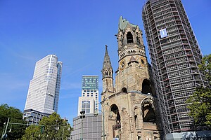 Die Kaiser-Wilhelm-Gedächtniskirche in Berlin mit ihrer markanten Ruine ist von modernen Hochhäusern umgeben. Rechts ist der Kirchturm eingerüstet. Links steht das auffällige weiße Hochhaus „Upper West“ mit seiner geschwungenen Architektur. Im Vordergrund sind Bäume zu sehen, die das Ensemble umrahmen.