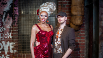 Ein Mann steht neben der Wachsfigur von RuPaul im Madame Tussauds. RuPaul trägt ein auffälliges rotes Glitzerkleid und großen Ohrschmuck, während der Mann eine Bluse mit Blumenmuster und eine Kappe trägt.