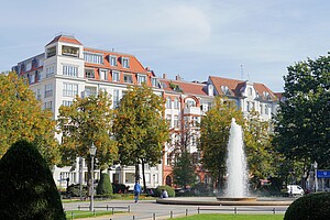 Ein majestätischer Springbrunnen in der Mitte des Viktoria-Luise-Platzes in Berlin. Ringsherum sind historische Gebäude mit roten Dächern zu sehen. Der Platz ist von Bäumen und gepflegten Grünflächen umgeben.