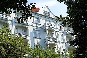 Ein weißer Altbau im Berliner Kollwitzkiez mit geschwungenem Giebel. Der Bau ist verziert und hat mehrere Balkone. Bäume umrahmen das Gebäude und die Blätter scheinen leicht im Wind zu wehen. Ein charmantes Beispiel Berliner Architektur aus der Gründerzeit.