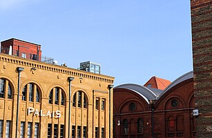 Das Foto zeigt eine Nahaufnahme von zwei Backsteingebäuden der Kulturbrauerei in Berlin. Auf dem einen Gebäude steht "PALAIS" in weißen Buchstaben, daneben sind bogenförmige Fenster und Dachkonstruktionen zu sehen. Der Himmel ist strahlend blau.