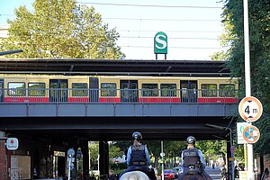 Eine Berliner S-Bahn fährt auf einer erhöhten Brücke mit einem grünen „S“-Schild im Hintergrund. Im Vordergrund stehen zwei berittene Polizeibeamte, die in die Szene hineinschauen.