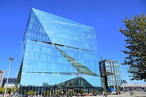 Ein modernes, gläsernes Gebäude in dreieckiger Form reflektiert den strahlend blauen Himmel. Die Fassade hat geometrische Muster, die wie ein Kreuz erscheinen. Links ist eine Straßenlaterne, rechts die Architektur des Hauptbahnhofs.