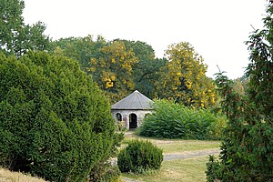 Ein kleiner, steinerner Pavillon steht zwischen dichten Büschen und hohen Bäumen, deren Blätter herbstliche Farben annehmen. Der Himmel ist bedeckt, aber die Szene wirkt friedlich und ruhig.