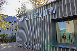 Das moderne Stadtbad Tiergarten in Berlin mit geschwungener, wellenartiger Fassade. Die Herbstsonne strahlt durch die Bäume, die das Gebäude umgeben.