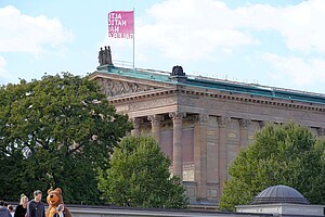 Die Alte Nationalgalerie auf der Berliner Museumsinsel, im Hintergrund das historische Gebäude mit Säulen und einem prächtigen Giebel. Im Vordergrund ein verkleideter Berliner Bär, umgeben von Passanten, während Bäume das Gebäude teilweise verdecken.