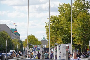 Eine belebte Szene auf der Straße Unter den Linden, mit Menschen, die zwischen Bauzäunen flanieren. In der Ferne sieht man das Brandenburger Tor, eingerahmt von Bäumen und deutschen Flaggen.