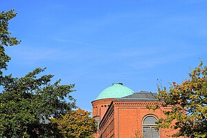 Die historische Fassade der Justizvollzugsanstalt Moabit mit ihrer roten Backsteinarchitektur und dem grünen Kuppeldach, eingerahmt von Bäumen und blauem Himmel.