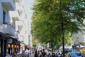 Eine belebte Berliner Straße, gesäumt von Altbauhäusern mit weißen Fassaden. Links im Bild befinden sich Restaurants und Cafés mit Außenbereichen, in denen viele Menschen sitzen. Rechts steht eine dichte Baumreihe, die Schatten auf die Straße wirft. Fußgänger und Radfahrer beleben die Szene.