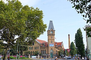 Das Foto zeigt den Hauptturm der Kulturbrauerei in Berlin mit seinem charakteristischen spitzen Dach und roten Backsteinmauern. Im Vordergrund sind Fußgänger zu sehen, die die Straße überqueren, während links ein grüner Baum das Bild rahmt. Ein blauer Himmel sorgt für freundliche Stimmung.