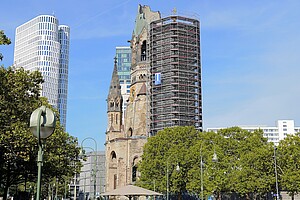 Der Kurfürstendamm in Berlin mit der Kaiser-Wilhelm-Gedächtniskirche im Hintergrund und moderner Bebauung im Vordergrund. Der Platz ist von Blumenbeeten und Sitzgelegenheiten umgeben.