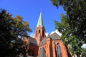 Der rote Backstein-Kirchturm einer Kirche mit einem hohen, grünen Dach aus Kupfer erhebt sich in den blauen Himmel. Der Turm ist von grünen Bäumen umgeben, die an den Rändern des Bildes sichtbar sind, und das Sonnenlicht taucht die Szenerie in ein helles, freundliches Licht. Die Architektur zeigt spitze Bögen und ein schlankes Design, typisch für neugotische Kirchen.